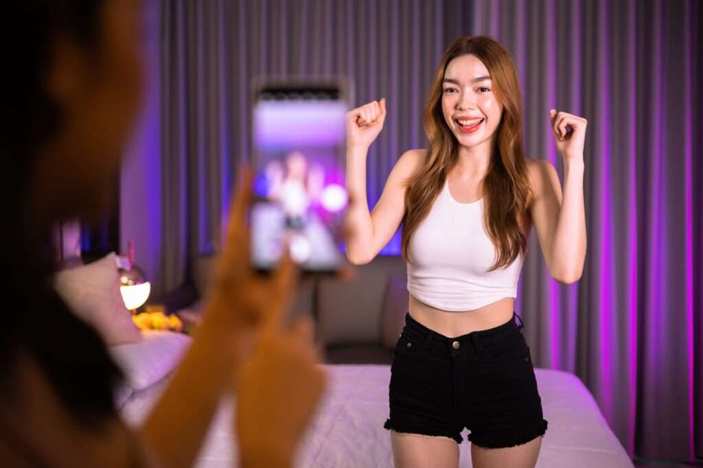 Young woman in crop top and shorts dancing for phone on bed, purple lights.