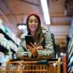 Happy woman using mobile app on her phone while shopping in supermarket.